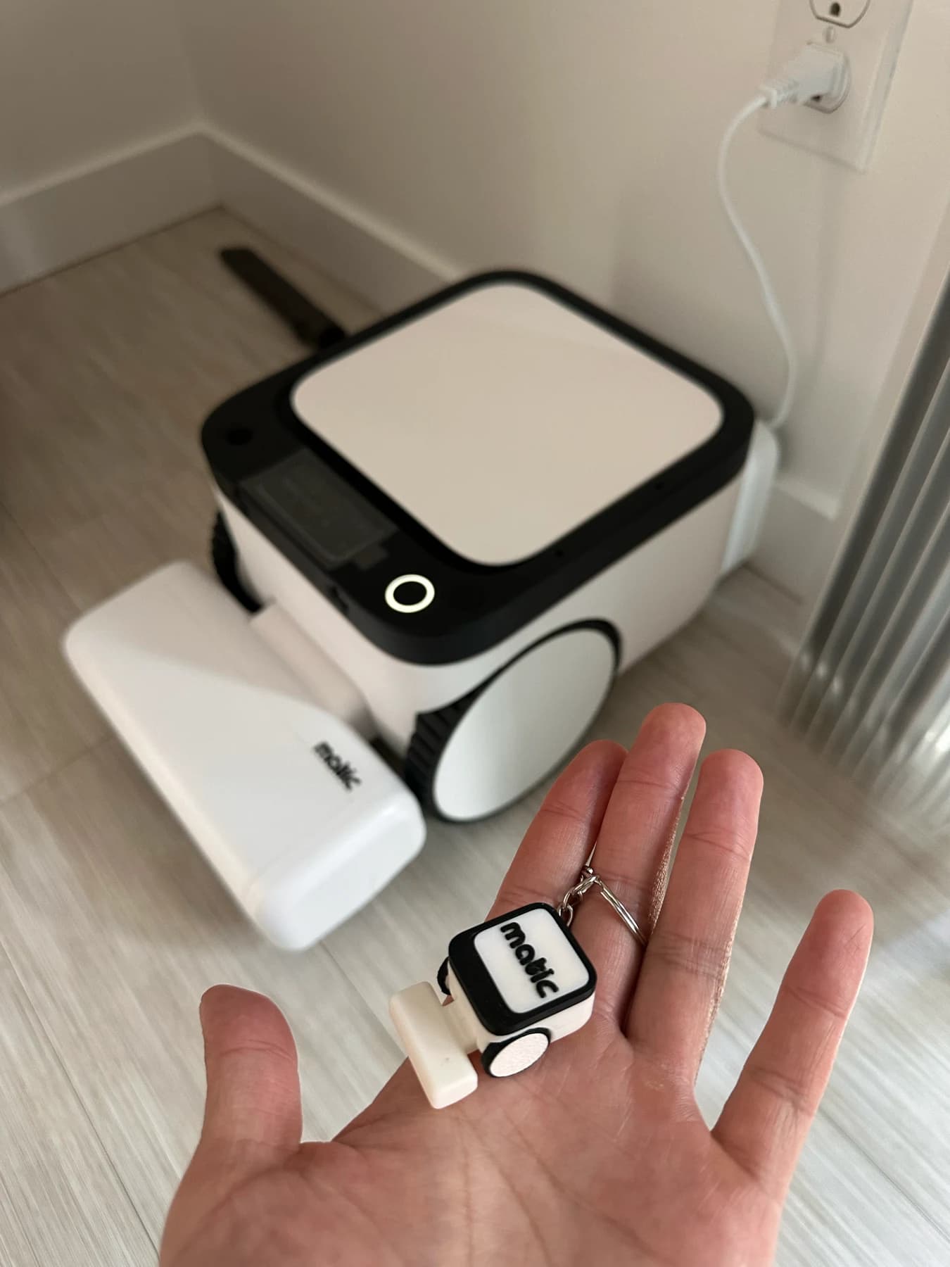 A hand holding a miniature Matic keychain, with the full-size robot parked behind it