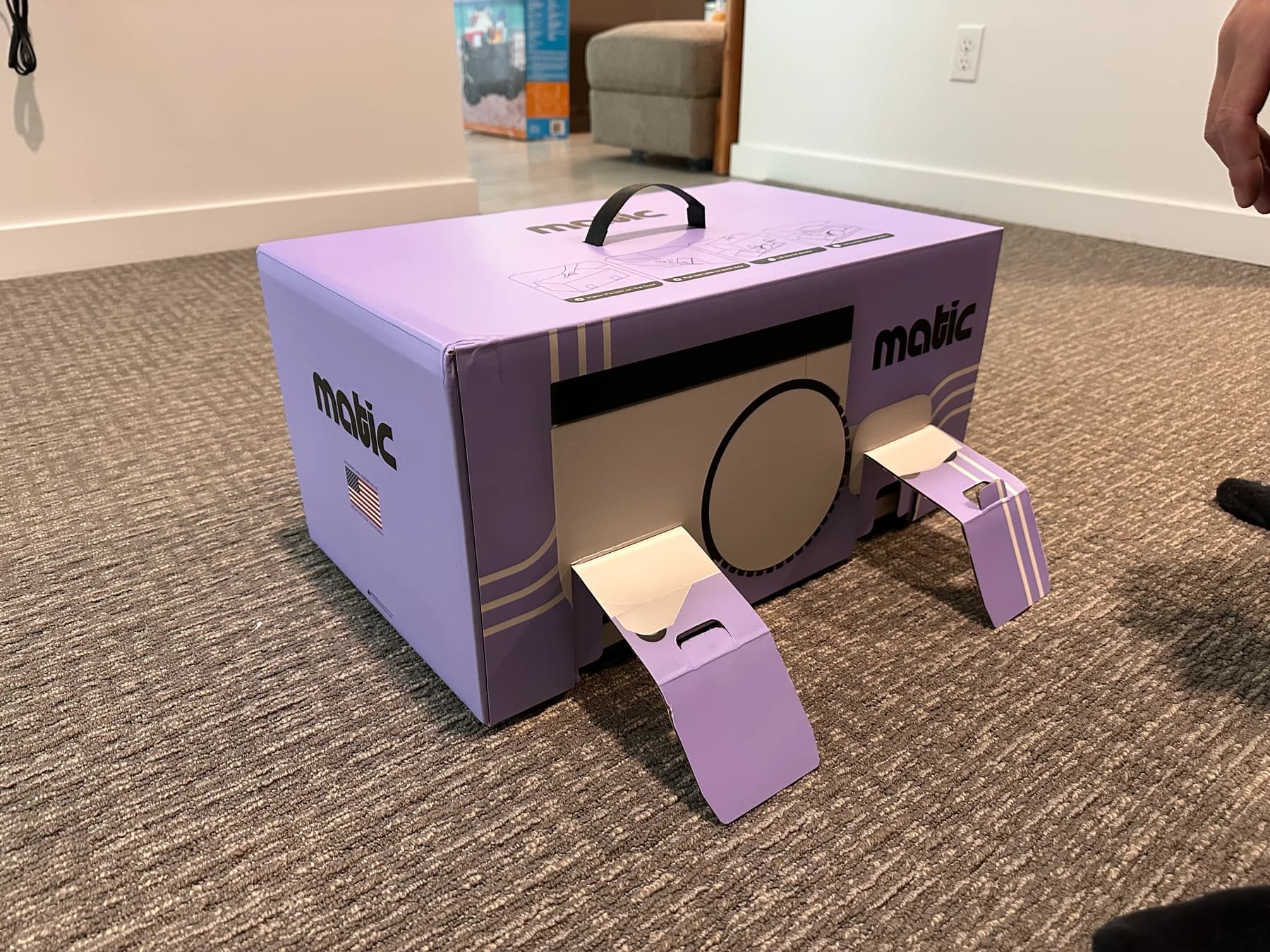 Matic's purple shipping box on carpet, with the two front cardboard tabs pulled open